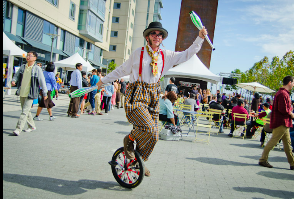 Slim juggling pins on a unicycle at an outdoor event in fun clown clothes