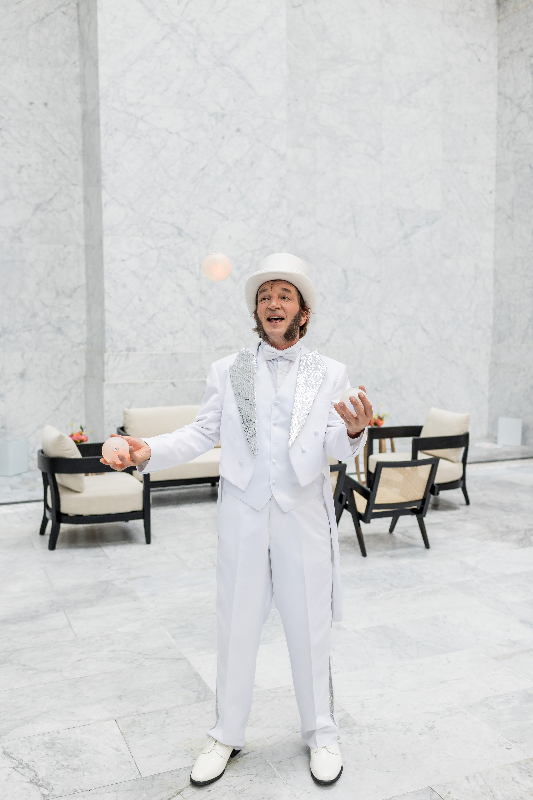 Slim juggling LED balls in a white suit and top hat in a white marble room