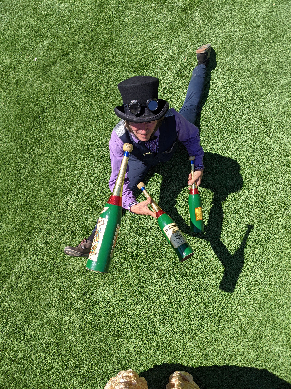Slim juggling bottles while doing a split on grass, wearing a top hat and steam punk goggles