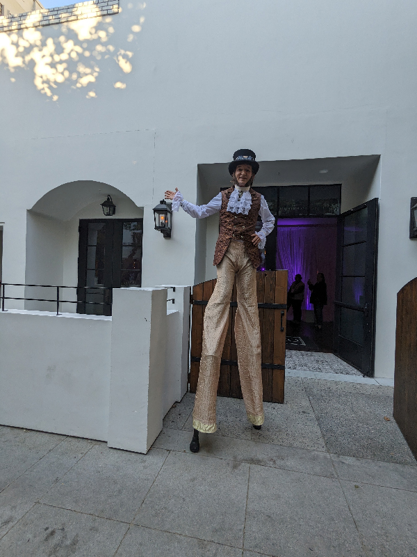Slim walking on stilts in a victorian steam punk outfit in front a of a venue with purple lights inside