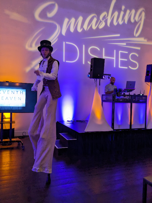 Slim dancing on stilts in victorian steam punk outfit, behind him is a DJ and a projection that says Smashing Dishes