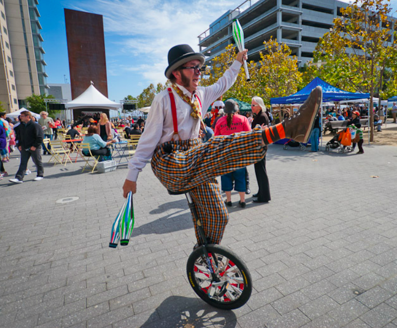 Slim juggling pins on a unicycle in an outdoor square with a stage and booths in the background and people milling around