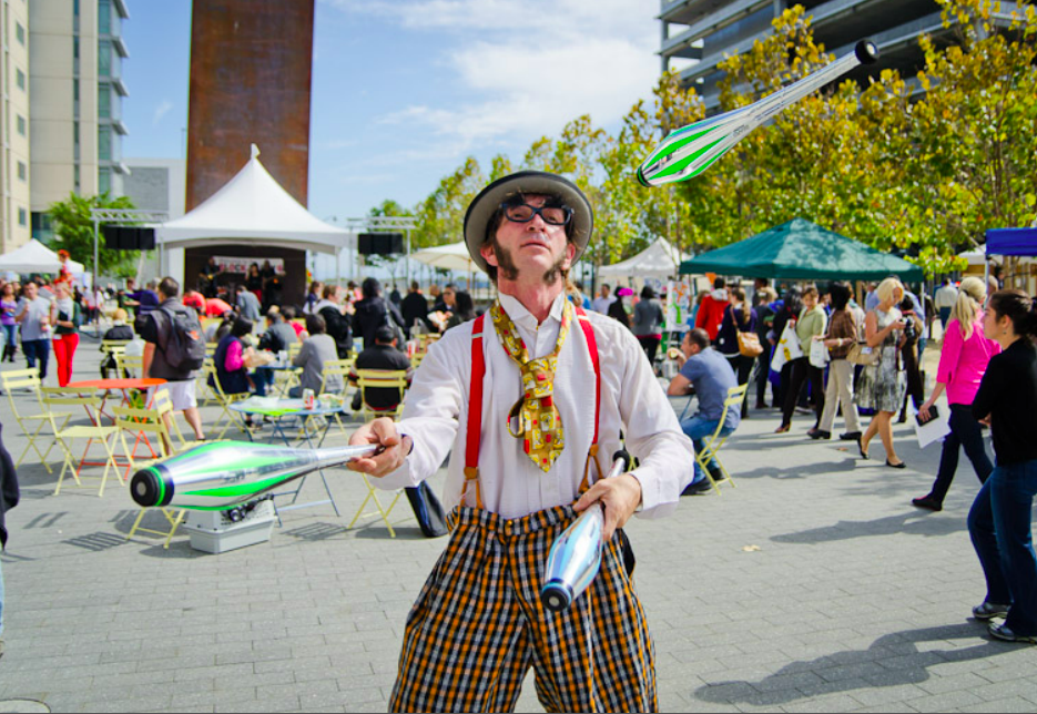 Slim juggling pins in an outdoor square wearing a colorful outfit with overalls and silly glasses