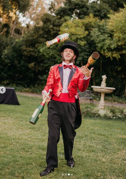 Slim juggling bottles in a garden area wearing a sparkly red entertainer's suit and top hat