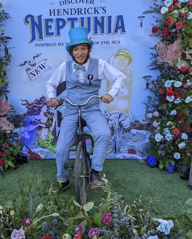 Slim posing for a photo on a bicycle in front a garden event backdrop wearing a blue old timey outfit